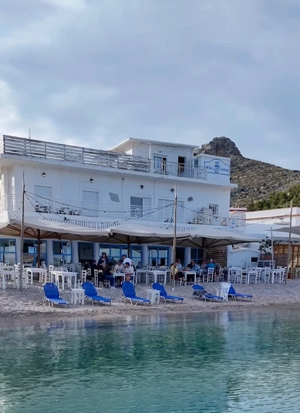 Leros'ta denize sıfır konumda, beyaz binası ve büyük şemsiyelerle plajda yer alan Psaropoula tavernasında insanlar masalarda yemek yerken, ön planda berrak denizin hemen yanında mavi şezlonglar görülüyor, arka planda ise kayalık bir dağ yükseliyor.