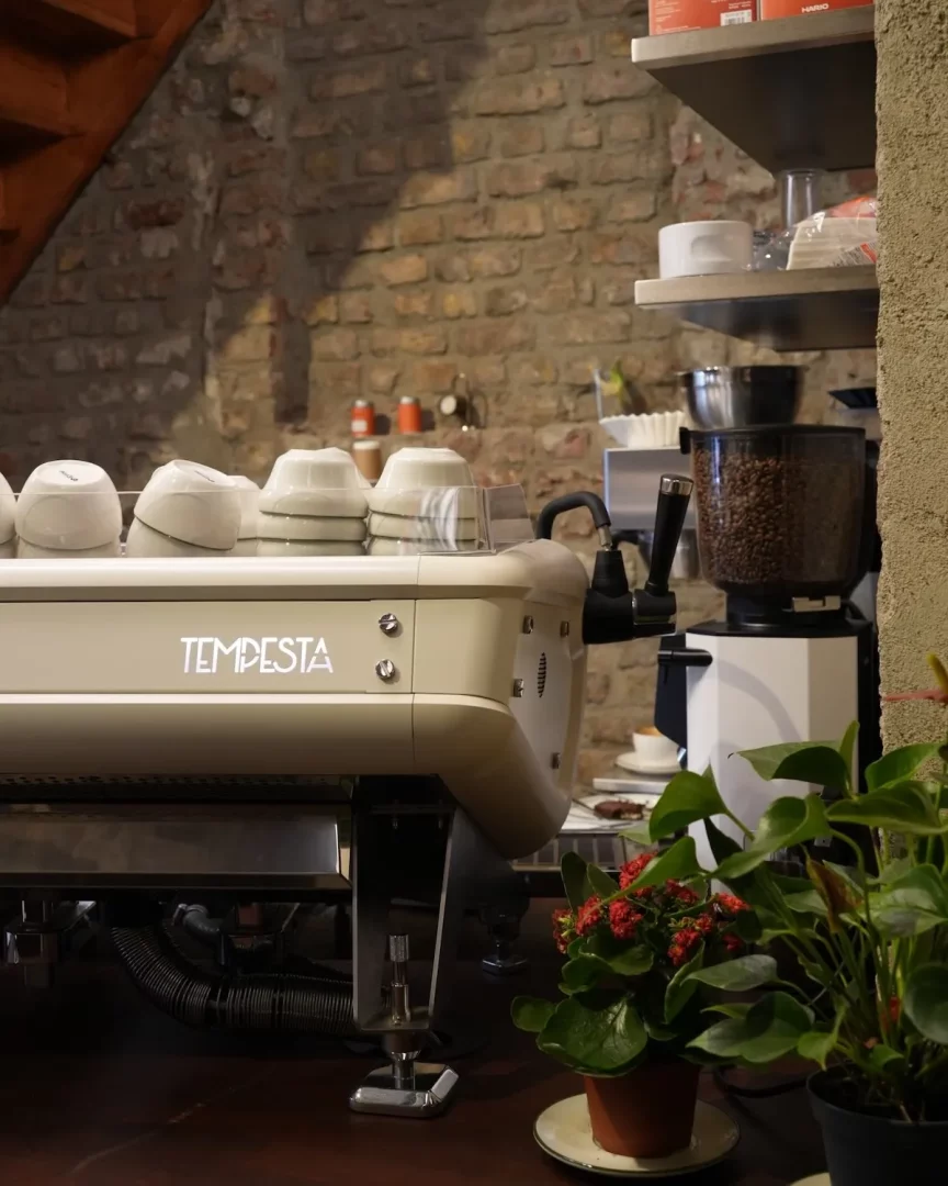 Bir kahve kavurma dükkanının içinden çekilmiş bu detaylı karede, üzerinde beyaz fincanların dizili olduğu bej renkli modern bir Tempesta espresso makinesi ve sağında kahve çekirdekleriyle dolu bir öğütücü, arkada ise rustik tuğla duvar önünde Arc Co. kahve deneyimine dair sıcak bir atmosfer sunuyor.