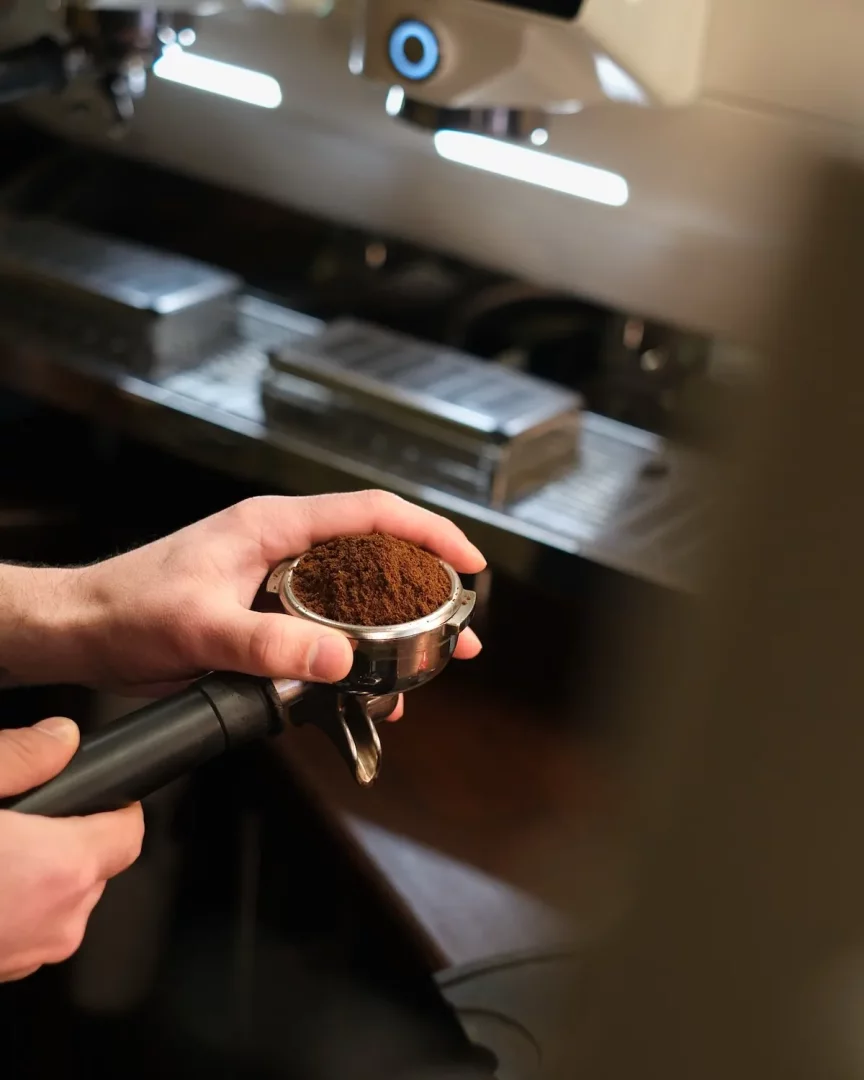 Profesyonel bir kahve kavurma işletmesinde veya kafede, bir barista espresso makinesinin önünde, mükemmel bir kahve demlemesi hazırlamak üzere parlak metalden yapılmış ve simsiyah saplı portafiltreyi taze, ince çekilmiş kahveyle doldurmuş bir şekilde iki eliyle nazikçe tutuyor.