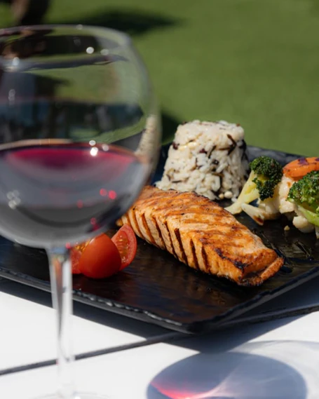 Argo Restaurant'ın açık hava masasında, parlak güneş altında, ızgara somon balığı, pilav, buharda pişirilmiş brokoli ve karnabahar gibi sebzeler ile kiraz domates dilimleri içeren lezzetli bir yemeğin yanında, ön planda bir kadeh kırmızı şarap bulunmaktadır.