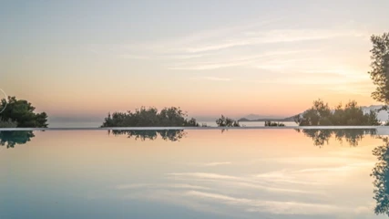 Armonia Bay Hotel'in sonsuzluk havuzunun cam gibi durgun yüzeyinde, denizin üzerinde batan güneşin pastel turuncu ve pembe tonlarındaki gökyüzü, uzak adalar ve kıyıdaki yemyeşil ağaçlar berrak bir şekilde yansıyor, huzurlu bir atmosfer yaratıyor.