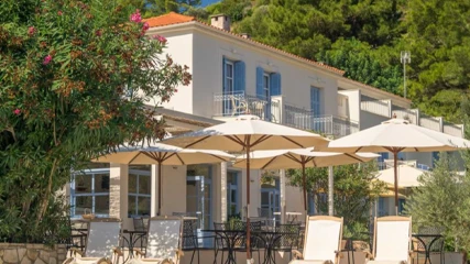 Armonia Bay Hotel'in bej güneş şemsiyeleri altındaki siyah masa, sandalyeler ve şezlonglarla donatılmış yemyeşil ağaçlar ve çiçekli çalılarla çevrili açık hava dinlenme alanı, açık mavi panjurlu ve balkonlu beyaz binanın Akdeniz tarzı kiremit çatısı ve zeytin ağaçlarıyla bütünleşerek huzurlu bir görünüm sunuyor.