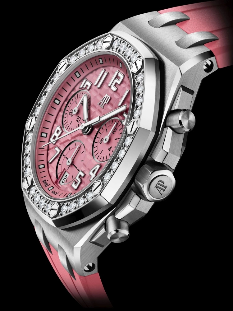 Siyah arka plan üzerinde, açılı bir perspektiften görünen bu Audemars Piguet Royal Oak Offshore lüks kronograf kol saati, pembe dokulu kadranı, pırlanta işlemeli sekizgen bezeli ve pembe kauçuk kordonuyla göz alıcı bir şıklık sunuyor.