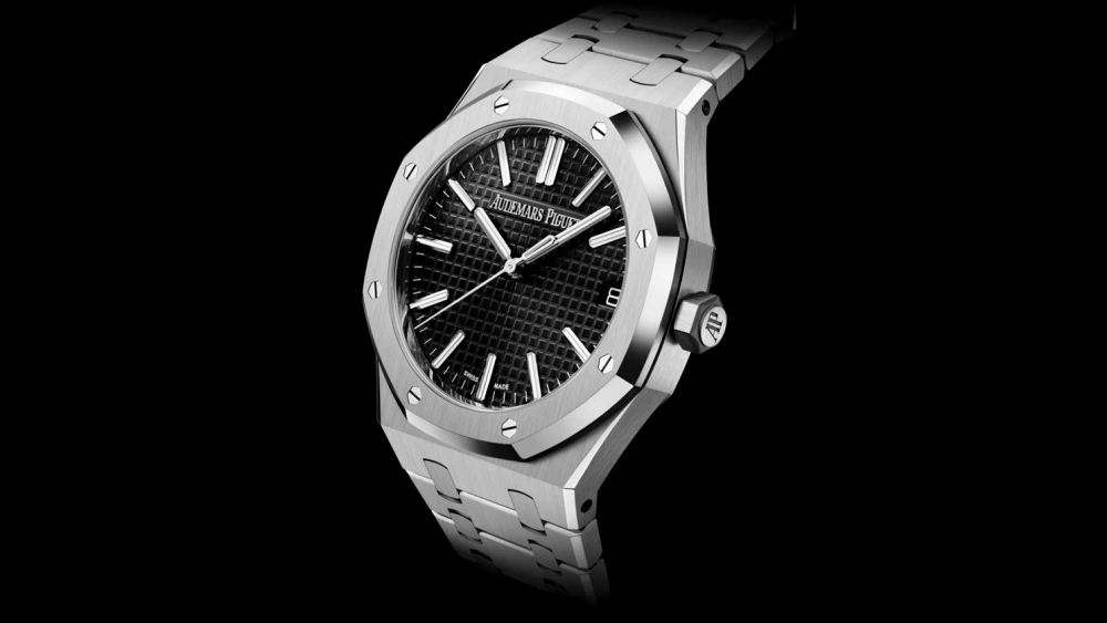 Audemars Piguet Royal Oak modelinin fırçalanmış paslanmaz çelik kasası ve entegre bileziğiyle birlikte, karakteristik Grande Tapisserie desenli siyah kadranı, beyaz baton indeksleri, akrep ve yelkovanı, saat 3 yönündeki tarih penceresiyle siyah bir fon üzerinde açılı bir şekilde sergilenen detaylı bir görünüm.