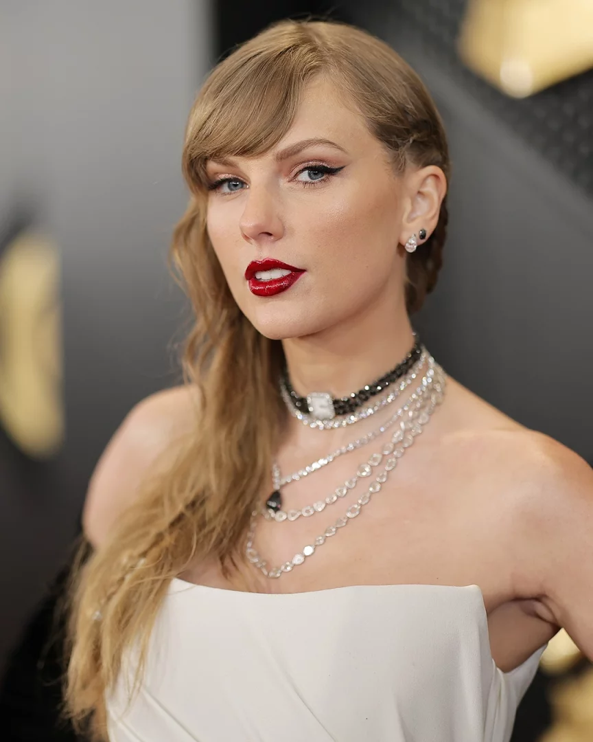 Bal sarısı saç rengine sahip Taylor Swift'in perçemli ve dalgalı saçlarıyla, göz alıcı kırmızı ruju, kuyruklu eyelinerı, pırlanta küpeleri ve straplez beyaz elbisesi üzerine taktığı katmanlı kolyeleriyle çekilmiş yakın plan portresi.
