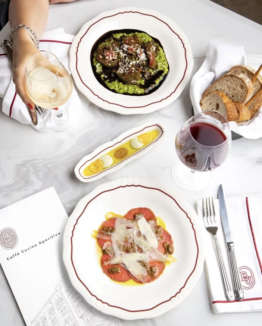 Beyaz mermer bir masanın üzerinde, Banca Unione Karaköy'deki İtalyan mutfağının enfes örneklerini sergileyen zengin bir yemek sofrası; parmesan peynirli carpaccio, bol soslu risotto, dilimlenmiş ekmek, bir kadeh beyaz şarap tutan bir el ve bir kadeh kırmızı şarap ile markalı menü ve peçeteler özenle düzenlenmiş.