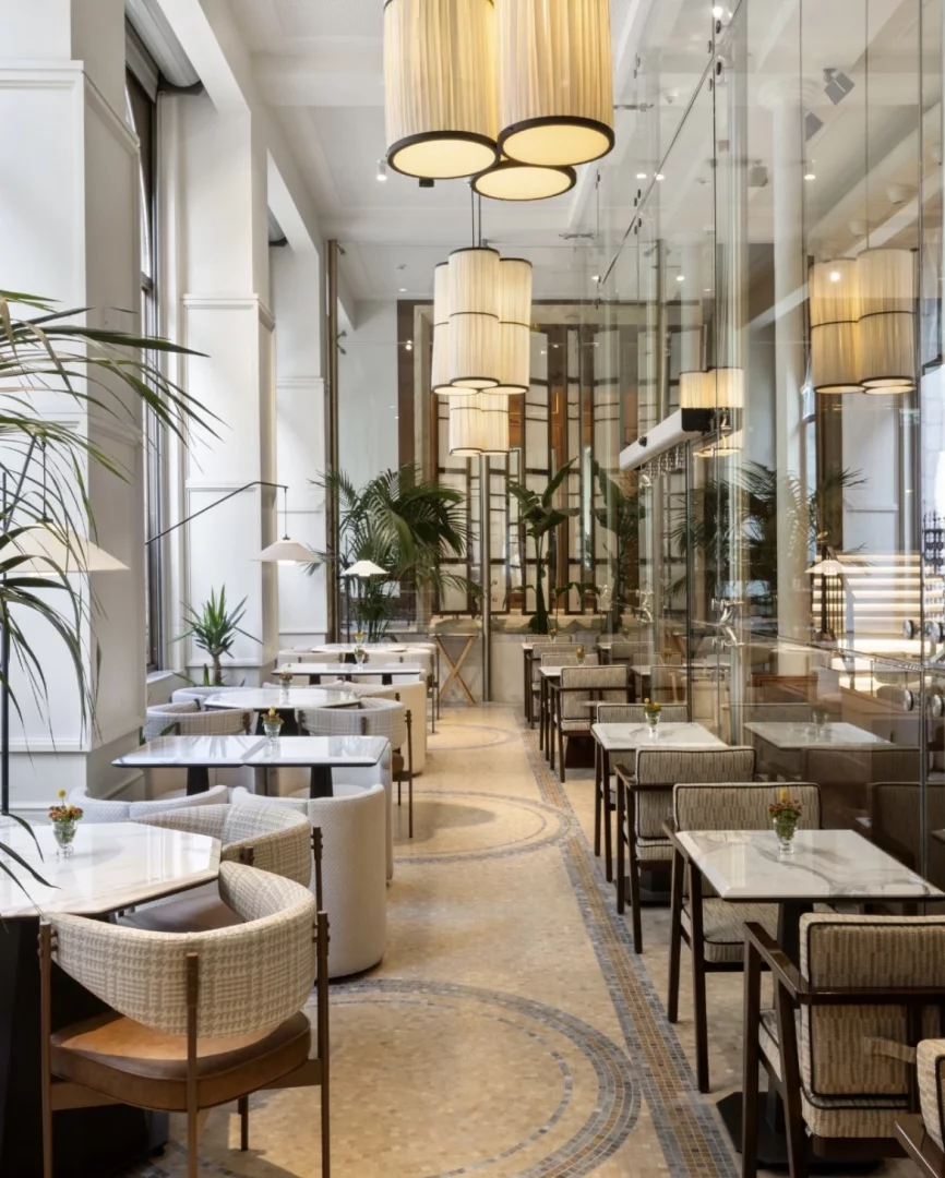 Geniş pencerelerden gelen doğal ışıkla aydınlanan, şık tasarımlı bir restoran veya kafe iç mekanı; mozaik desenli zeminde sıralanmış beyaz mermerli masalar, hasır dokulu sandalyeler, uzun, pilili sarkıt lambalar ve ferah bir atmosfer yaratan büyük salon bitkileri dikkat çekiyor.