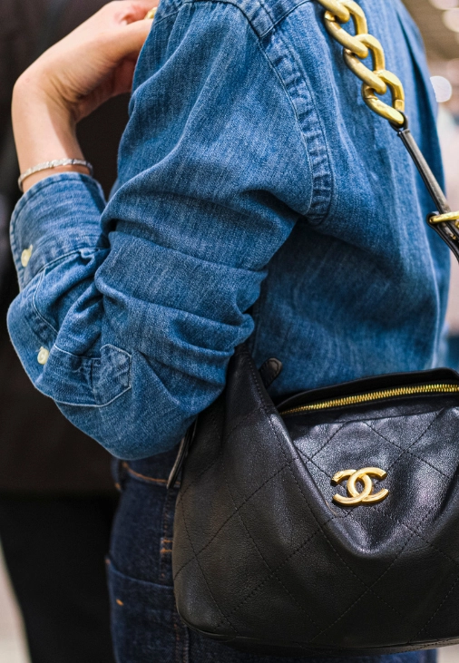 Mavi kot gömlek giymiş, kolunda ince bir gümüş bileklikle süslenmiş bir kişinin omzunda, ön yüzünde belirgin altın rengi Chanel logosu, kapitone deri deseni ve kalın altın zincir askısı bulunan, şık siyah bir çanta, yeni lüksün zamansız zanaat ve tasarım odaklı dönüşümünü sembolize ediyor.