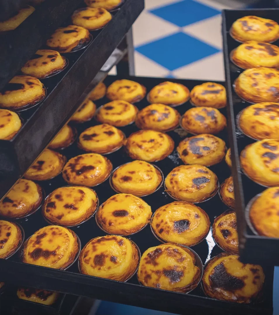 Portekiz'in meşhur yumurtalı tartları, altın rengi kabukları ve hafifçe karamelize olmuş üst kısımlarındaki koyu lekelerle dolu tepsilerde yan yana sıralanmış, taze pişmiş Pastel de Belém'ler ziyaretçilerini bekliyor.