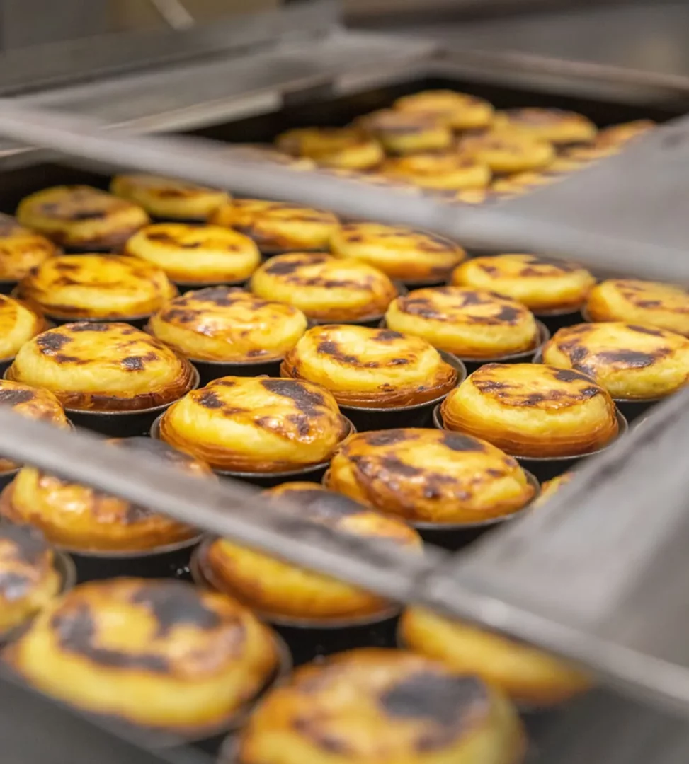 Fırın raflarında sıcacık bekleyen, üstleri karamelize olmuş ve hafifçe yanık izleri taşıyan onlarca geleneksel Portekiz kremalı tartı, yani pastel de nata, göz alıcı bir şekilde sıralanmış.