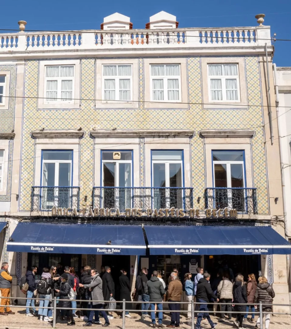 Belém'deki Antiga Fábrica dos Pastéis de Belém'in mavi-beyaz çinilerle süslü tarihi binasının önünde, Pastéis de Belém tatlılarını almak için uzun kuyruk oluşturan kalabalık insanların canlı bir görüntüsü, mekanın popülerliğini ve geleneksel lezzetini vurguluyor.