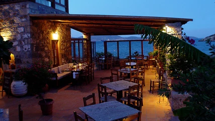 Patmos'taki Benetos Restaurant'ın taş mimarisi ve ahşap pergolasıyla bezenmiş, deniz kenarındaki dış mekanı, akşamüstü aydınlatmasıyla zarif masaları ve Ege'nin dingin sularını gözler önüne sererek romantik bir akşam yemeği atmosferi sunuyor.