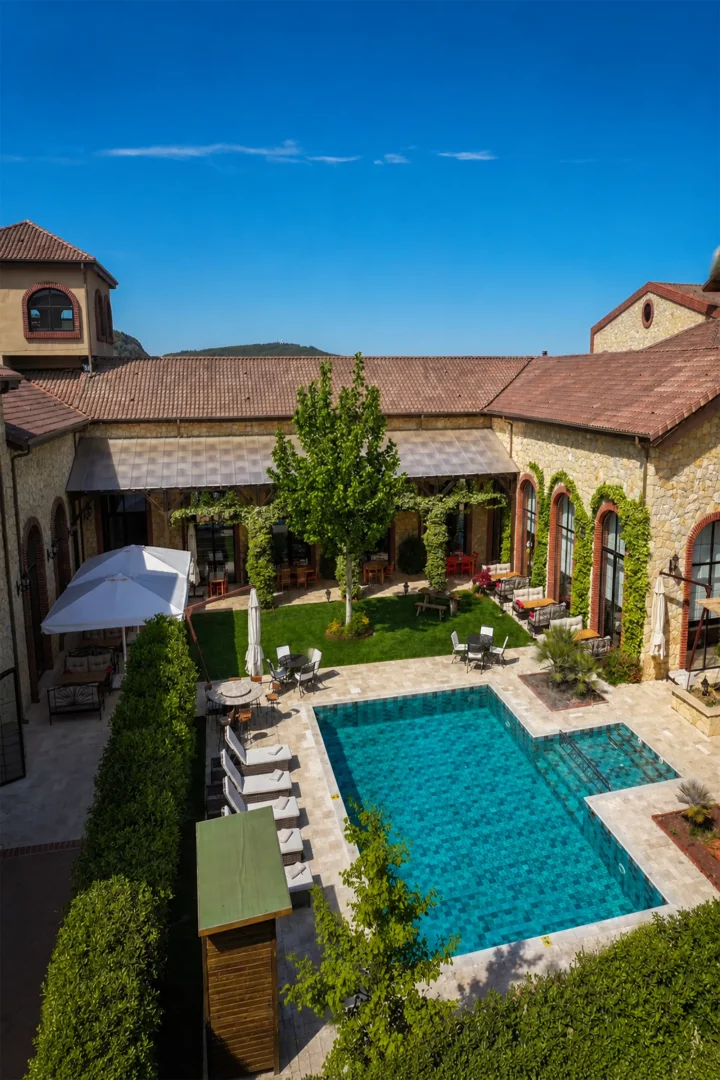 Çanakkale'de bulunan Bengodi Boutique Hotel'in kuşbakışı görüntüsünde, kiremit çatılı taş yapılarla çevrili yemyeşil bir bahçe içerisinde, güneşli mavi gökyüzü altında berrak mavi mozaik karolarıyla dikkat çeken bir açık yüzme havuzu ve etrafında şezlonglar ile zarif dış mekan oturma alanları görülüyor.