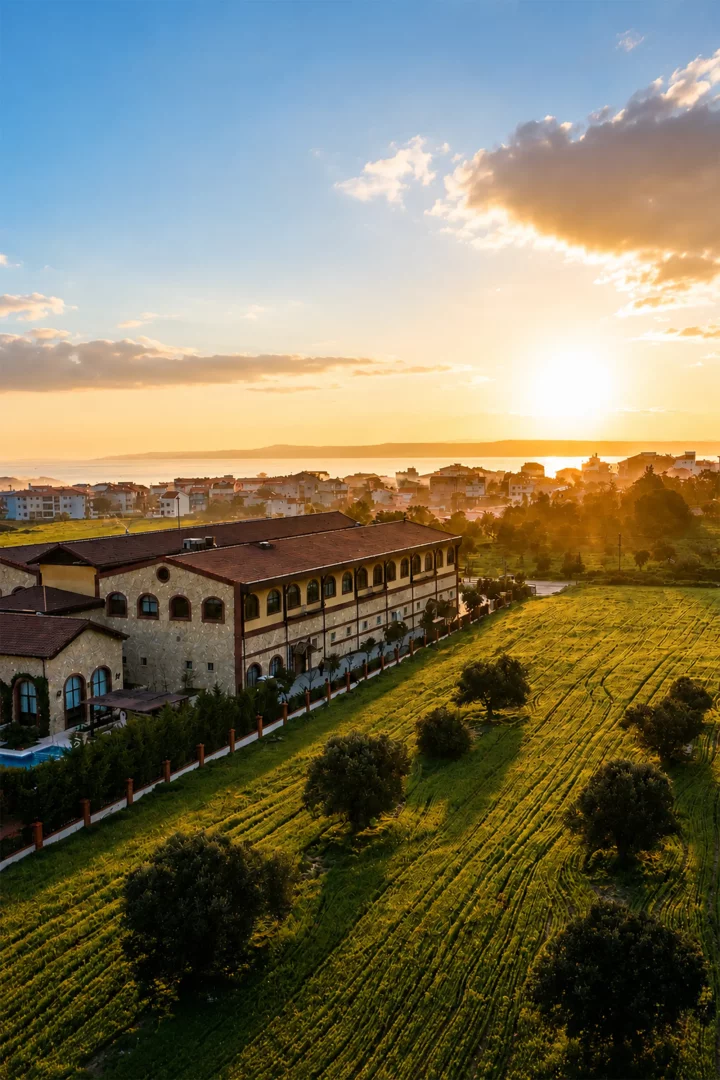 Gün batımının altın rengi ışıkları altında, Bengodi Boutique Otel'in kahverengi çatılı, taş detaylı binası, önündeki denize uzanan izli yeşil tarlalar ve uzaktaki sahil kasabasıyla birlikte huzurlu bir manzarayı gözler önüne seriyor.