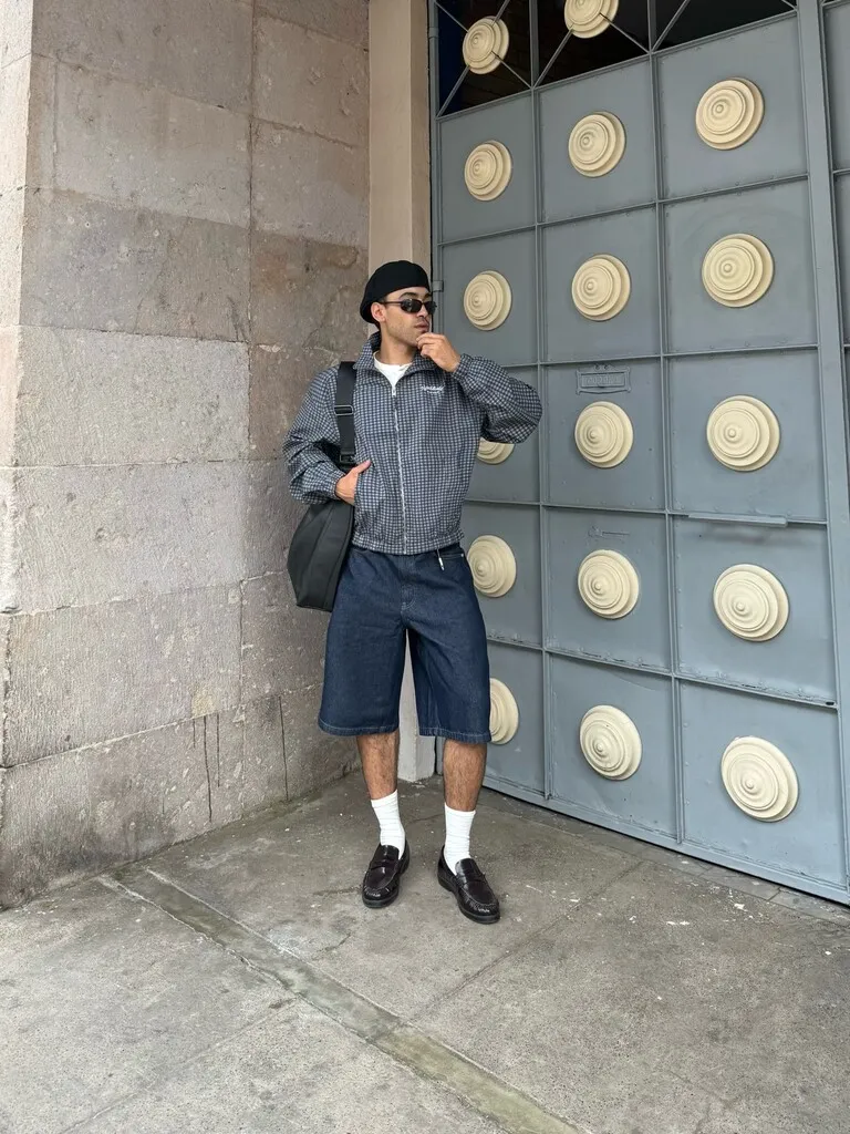 Bir erkek model, siyah bere, güneş gözlüğü, ekose ceket ve özellikle bol kesim koyu renk denim şortuyla gri taş duvar ve yuvarlak desenli metal bir kapının önünde poz vererek Bershka'nın yazlık stilini sergiliyor.