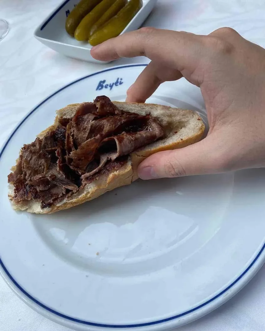 Beyaz bir masada, üzerinde ince dilimlenmiş ve nefis görünen döner kebabı ile dolu ekmek arası döner tutan bir el, mavi kenarlı Beyti restoran tabağının merkezinde belirgin; arka planda hafifçe bulanık turşular da iştah açıcı bir şekilde duruyor.