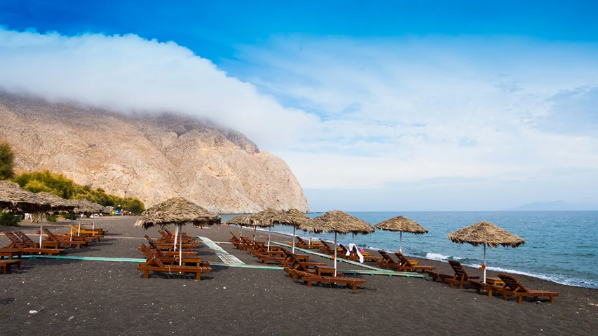Santorini'nin ünlü kara plajlarından birinde, bulutlarla çevrili sarp kayalıkların ve masmavi denizin önünde uzanan, gölgeli hasır şemsiyelerin altında düzenli sıralar halinde dizilmiş ahşap şezlonglar, sakin bir tatil manzarasını gözler önüne seriyor.