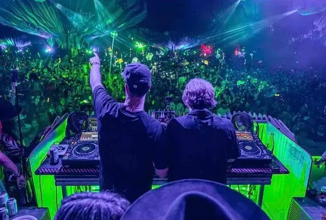Bob Moses'ın Club Set performansı sırasında, yeşil ve mor ışıklarla aydınlanmış DJ kabininin önünde elleri havada dans eden coşkulu bir kalabalıkla dolu, Afterglow elektronik müzik festivali atmosferi.