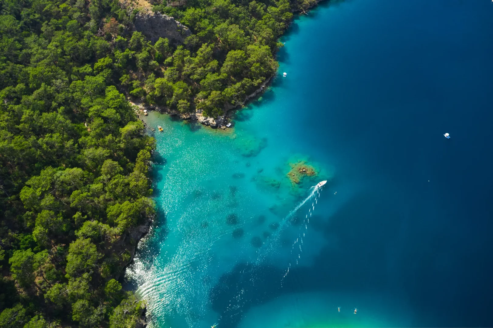 Bodrum ve Gökova'nın büyüleyici koylarını anlatan bu hava fotoğrafında, yemyeşil çam ormanlarıyla çevrili kıyının berrak turkuaz sularında ilerleyen küçük bir tekne, ardında uzun bir köpük izi bırakarak huzurlu bir mavi yolculuk anını gözler önüne seriyor.