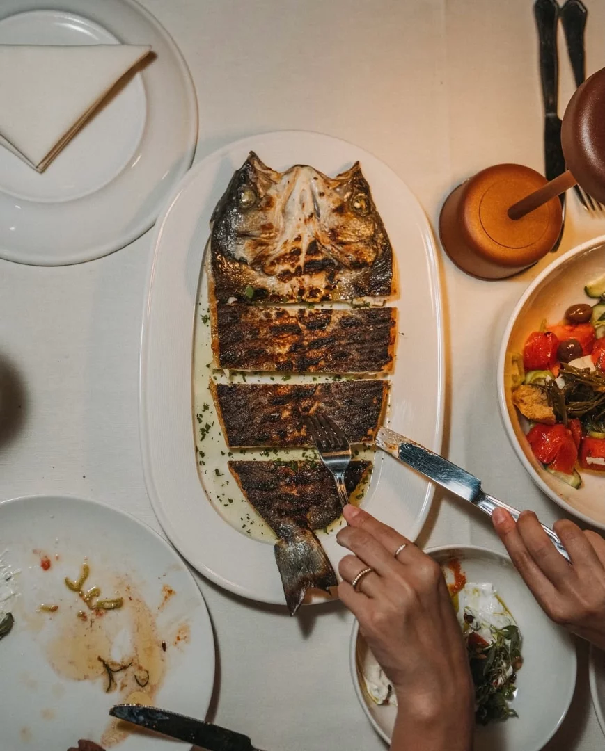 Bodrum'daki bir restoranda, beyaz sos ve yeşilliklerle sunulan, bütün olarak ızgaralanıp dilimlenmiş büyük bir balığın ana yemek olarak servis edildiği masada, bir kişinin çatal bıçakla yemeğe başladığı anı gösteren canlı bir an.