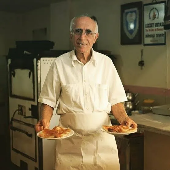 Beyaz gömlek ve önlük giymiş, gözlüklü ve güler yüzlü Tevfik Usta, arka planda geleneksel fırın ve Lezzet Ustası plaketinin asılı olduğu mutfak ortamında, her iki elinde de yeni pişmiş, altın rengi böreklerle dolu tabakları tutarak kameraya gülümsüyor.