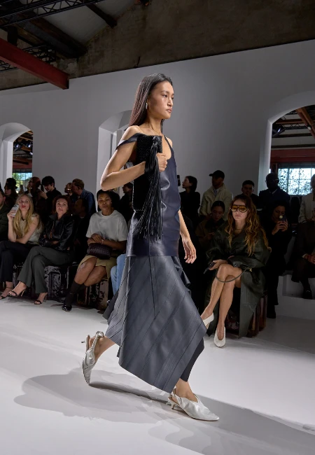 Bottega Veneta defilesinde podyumda yürüyen bir model, askısı omzundan düşmüş lacivert bir bluz, çapraz katman detaylarıyla hareketli ve yapısal asimetrik bir midi etek, açık renkli topuklu terlikler giymiş ve püsküllü bir çanta taşıyor, İlkbahar-Yaz 2026 asimetrik etekler trendini yansıtıyor.