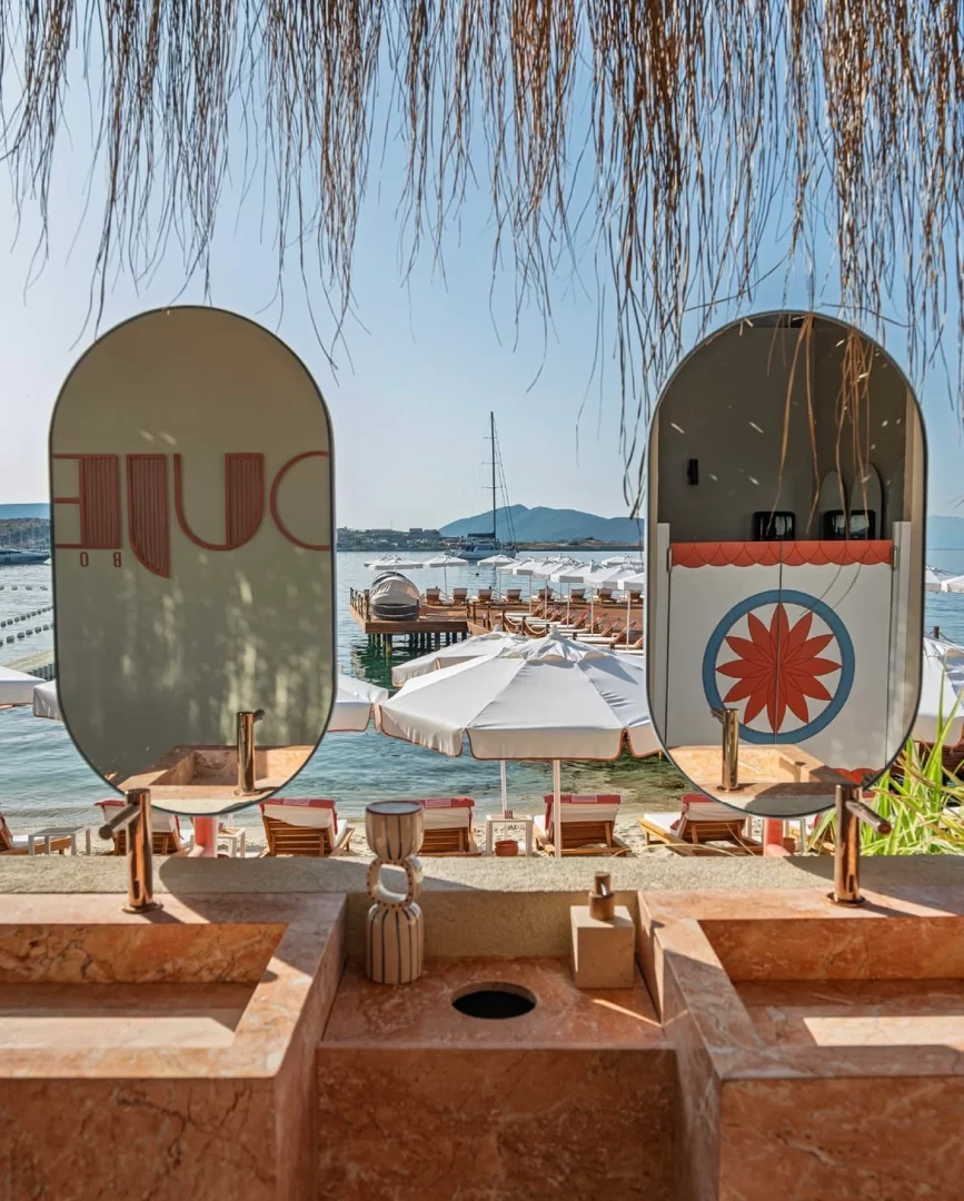 Bodrum'un gözde sahil mekanlarından birinde konumlanmış, mermer lavaboların ve aynaların ön planda olduğu lüks bir dış mekan tasarımı; aynalardan yansıyan şezlonglu iskele ve masmavi deniz manzarası ile hasır saçakların eşlik ettiği, özenli detaylara sahip bir alan sunuyor.