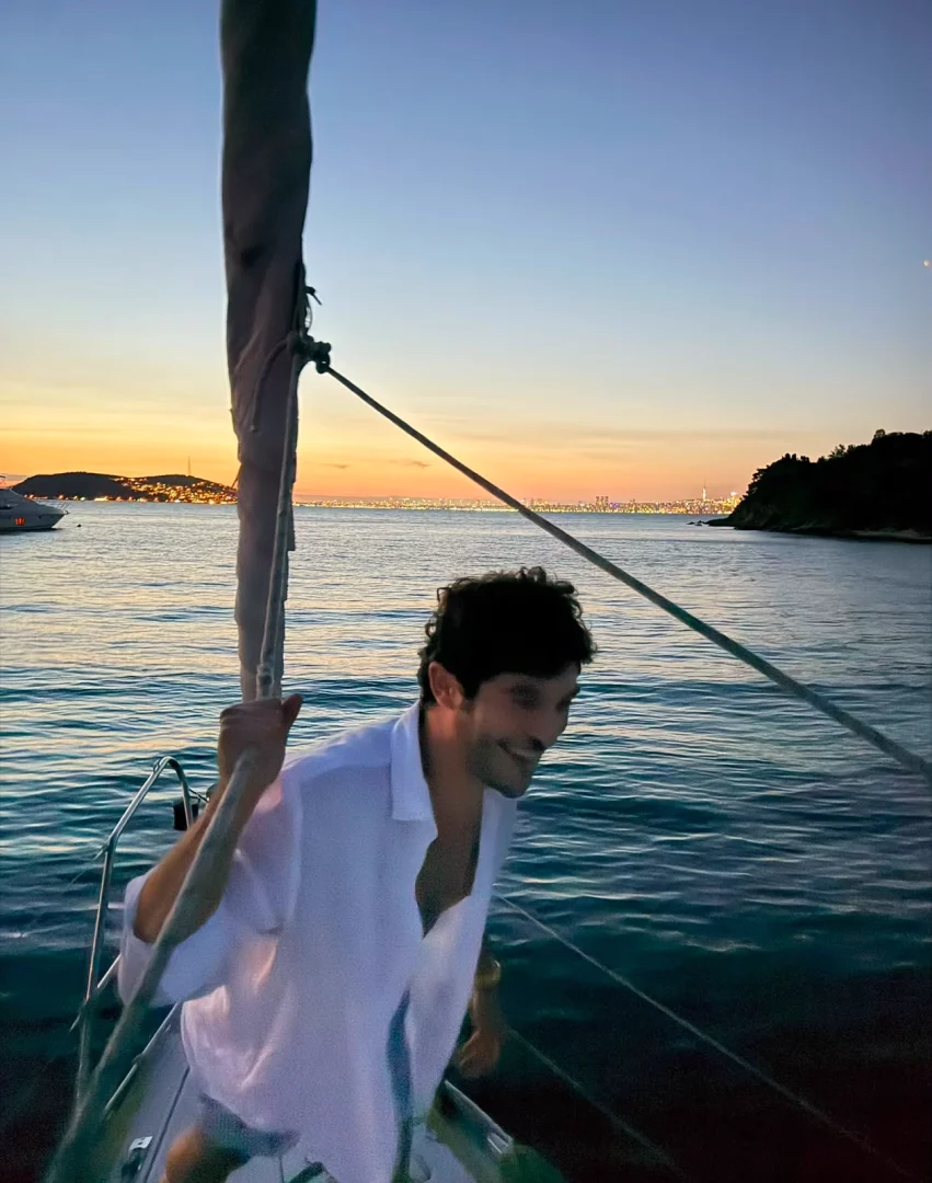 Oyuncu Burak Deniz, bir teknenin güvertesinde, gün batımının turuncu ve mor tonlarıyla aydınlanan denizin ve uzaktaki ışıklı şehir silüetinin huzurlu atmosferinde mavi yolculuğun keyfini çıkararak gülümserken görülüyor.