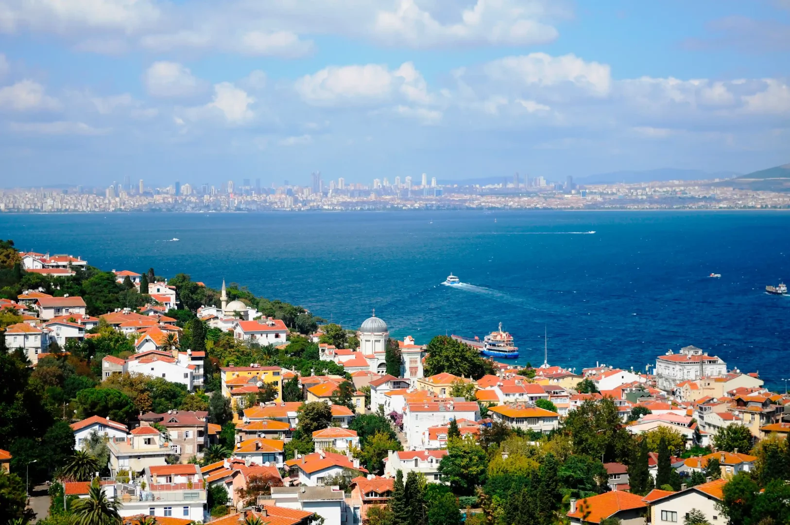Burgazada'nın yemyeşil yamaçlarında sıralanmış, kırmızı kiremitli çatılı evlerin masmavi Marmara Denizi ile buluştuğu panoramik manzarada, denizde ilerleyen tekneler ve ufukta yükselen İstanbul silüeti belirgin bir şekilde görülüyor.