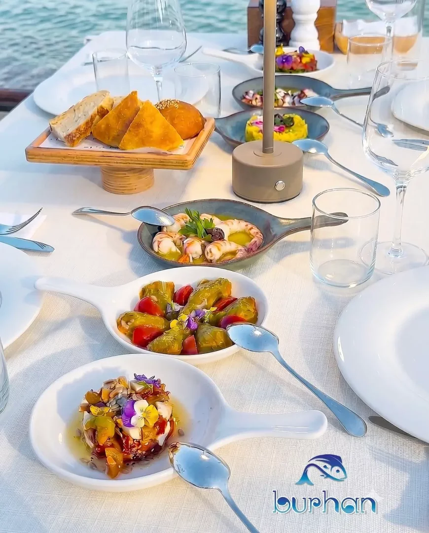 Bodrum'da, Burhan restoranının denize sıfır, beyaz örtülü zarif masasında, taze karidesli bir meze, biberli ve domatesli otlu bir başlangıç, yenilebilir çiçeklerle süslü peynirli bir tabak ve çeşitli ekmeklerin özenle sunulduğu zengin bir akşam yemeği.