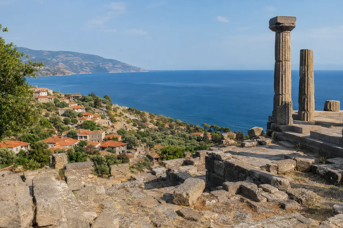Assos antik kentinden kalan iki büyük sütun ve taş kalıntılarının önde olduğu bu panoramik fotoğrafta, zeytin ağaçları arasında yamaç boyunca sıralanmış kırmızı çatılı köy evleri, masmavi Ege Denizi ve ufukta yükselen dağlar Çanakkale'deki bu önemli gezilecek yerin eşsiz güzelliğini gözler önüne seriyor.
