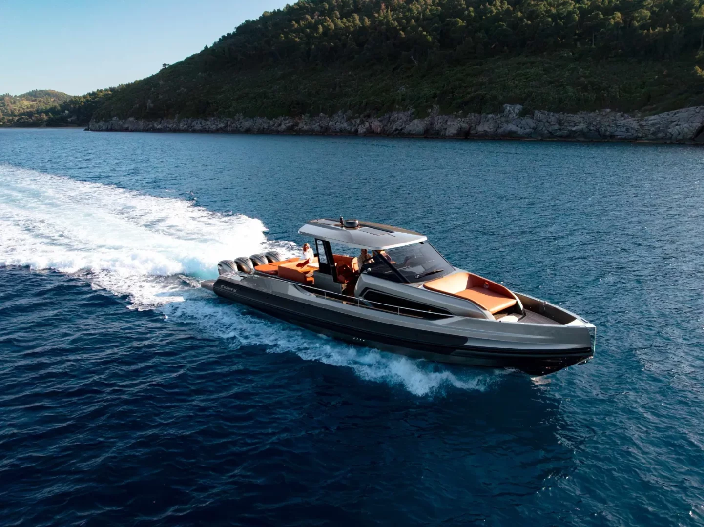 Bodrum açıklarında, yeşil tepelerin çevrelediği masmavi denizde hızla ilerleyen, gri gövdeli ve turuncu detaylı modern bir Cantieri Capelli Maxi RIB teknede iki kişi keyifli bir seyir yaparken, arkasında belirgin bir beyaz köpük izi bırakıyor.