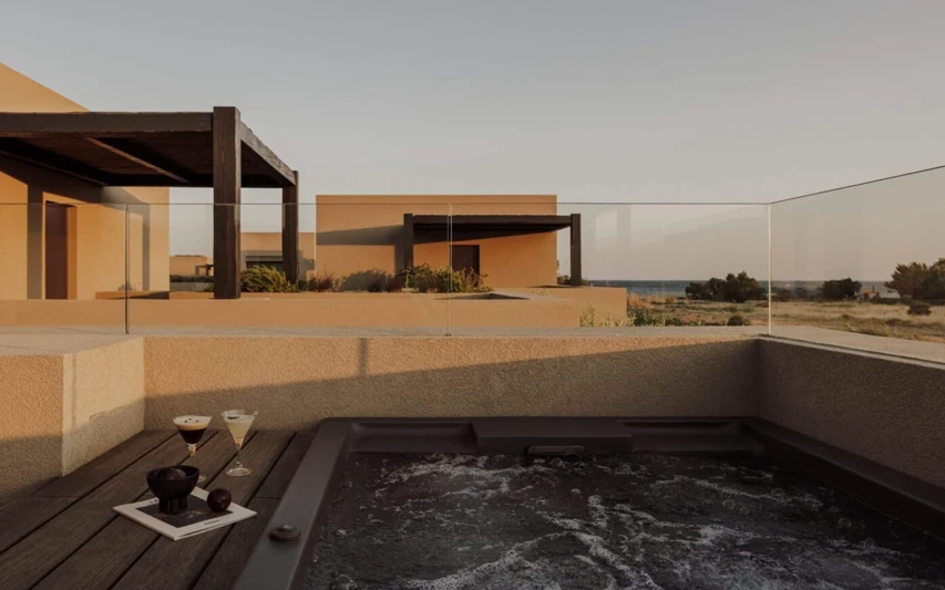 Casa Cook Samos otelinin minimalist tasarımlı jakuzili terasında, denize nazır modern binalar, koyu ahşap pergolalar, köpüren bir jakuzi, yanında iki kadeh kokteyl ve bir kitap ile huzurlu bir tatil dinlenme alanı gösteriliyor.