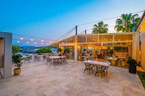 Akşamüstü maviliğinde, parlak ışıklarla aydınlatılmış ferah mermer terasta, denize nazır modern cam cepheli restoranın önünde, uzakta demirli teknelerin ve yemyeşil kıyıların görüldüğü, şık masa ve sandalyelerin misafirlerini beklediği keyifli bir açık hava mekanı.