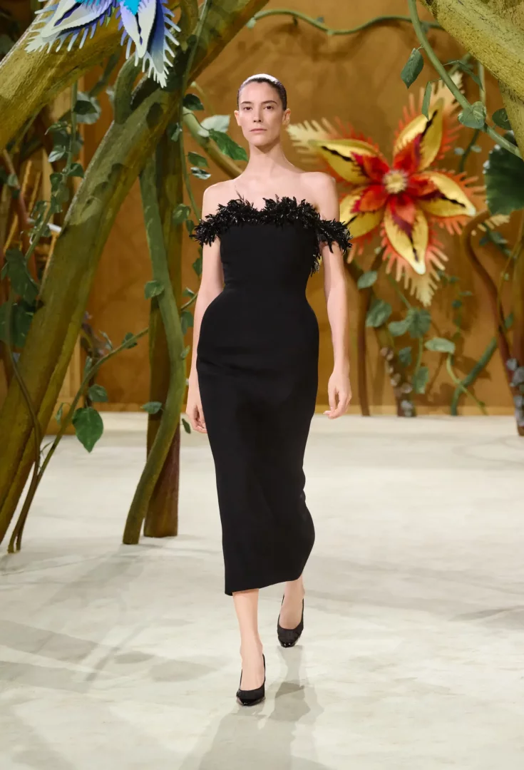 Omuzları siyah tüylü detaylarla süslenmiş straplez, siyah midi bir elbise giyen model, Chanel Sonbahar-Kış 2026 Couture Koleksiyonu defilesi için dev çiçekler ve sarmaşıklarla bezeli masalsı bir podyumda yürüyor.