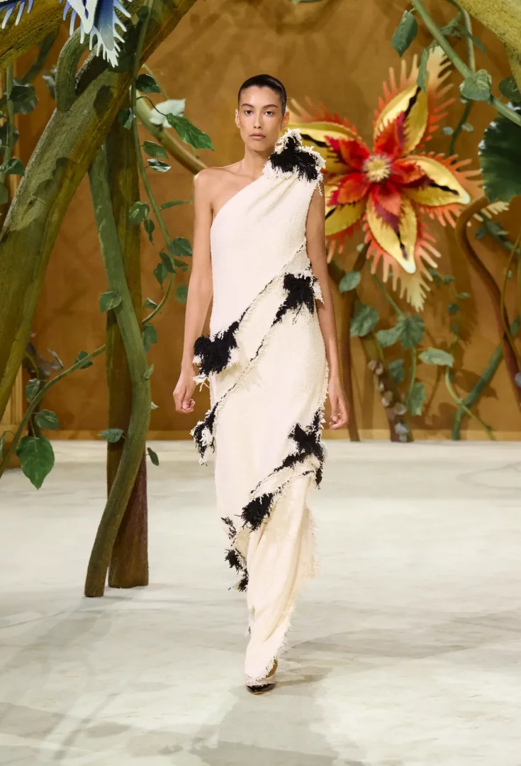 Paris Haute Couture Moda Haftası kapsamında sergilenen Chanel Sonbahar-Kış 2026 Couture koleksiyonundan bir model, masalsı bir atmosfer yaratan dev sarmaşıklar ve fantastik çiçeklerle çevrili podyumda, krem rengi, tek omuzlu, katmanlı ve siyah püsküllü detaylara sahip zarif bir elbiseyle ilerliyor.