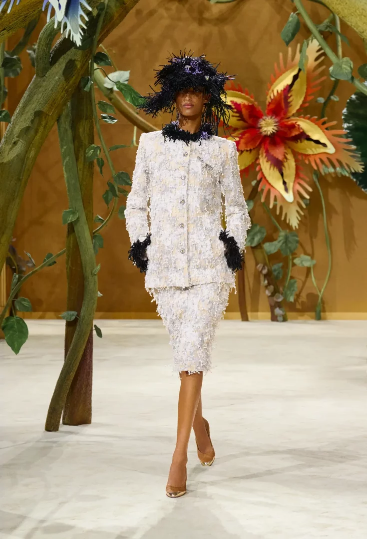 Chanel Sonbahar-Kış 2026 Couture Koleksiyonu defilesinde, masalsı bir podyumda yürüyen bir model, siyah tüylü yakalı ve manşetli, kabartmalı beyaz bir etek ceket takım ile kuş yuvasını anımsatan siyah bir şapka giyiyor.