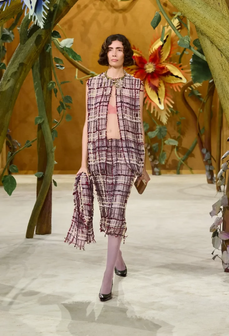 Chanel Sonbahar-Kış 2026 Couture Koleksiyonu'ndan bir görünüm sunan model, dev çiçekler ve bitki dallarıyla bezeli masalsı podyumda, pembe sütyen üzerine giydiği mor, krem ve bordo tonlarında, hafif ve püsküllü ekose tüvit yelek ile uyumlu diz altı eteği ve mor çoraplarıyla göz kamaştırıyor.