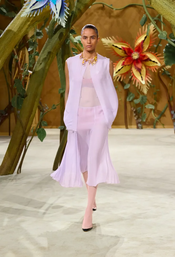 Paris Haute Couture Moda Haftası'nın masalsı temalı Chanel Sonbahar-Kış 2026 Couture koleksiyonundan bir model, dev çiçekler ve yapraklarla çevrili podyumda lavanta rengi şeffaf, hafif, pilili bir elbise ve gösterişli kolyesiyle yürüyor.
