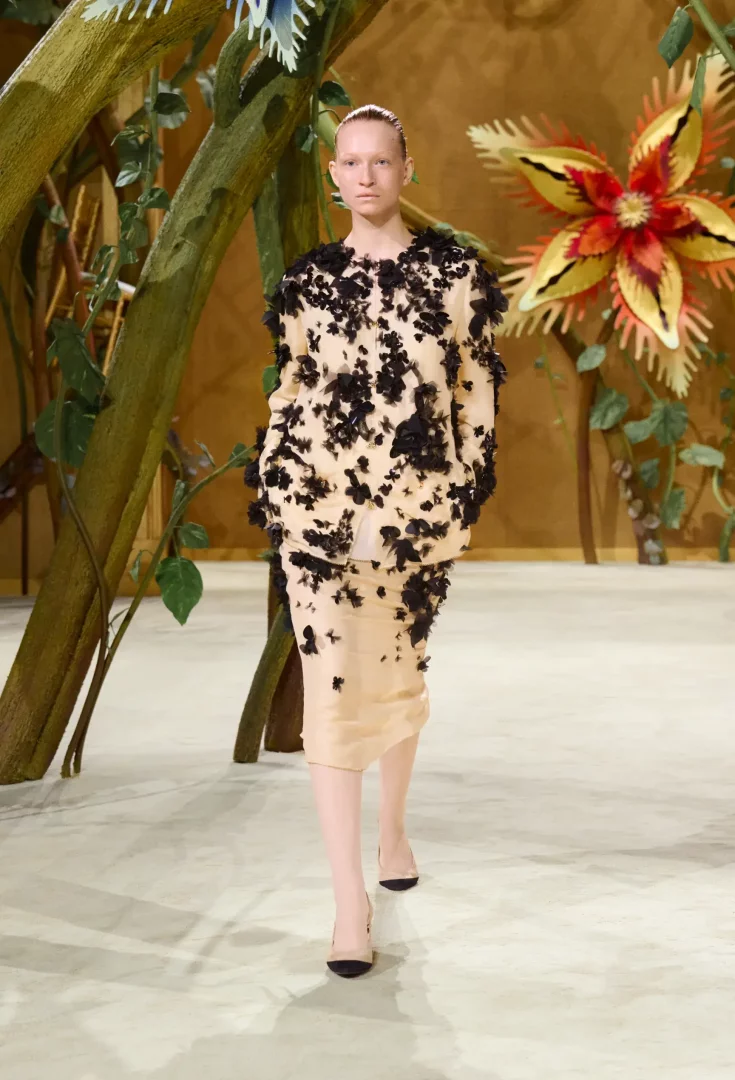 Chanel Sonbahar-Kış 2026 Couture koleksiyonundan, masalsı bir atmosferde, dev ağaç dalları ve egzotik çiçek figürleriyle süslenmiş podyumda, ten rengi bir ceket ve aynı tonlarda kalem etekten oluşan, tamamı siyah, üç boyutlu, kelebek benzeri işlemelerle kaplı özel bir parçayı sergileyen model.