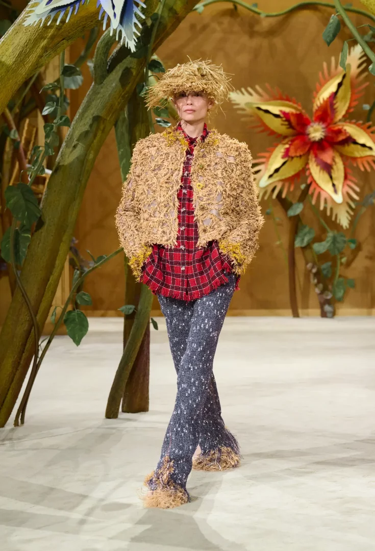 Chanel Sonbahar-Kış 2026 Couture Koleksiyonu defilesinde, devasa bitki ve çiçeklerle süslü masalsı podyumda yürüyen bir model; hasır görünümlü şapka, aynı dokuda kısa ceket, ekose gömlek ve paçaları püsküllü koyu renkli pantolonuyla hayal gücünü gündelik hayatla birleştiren bir görünüm sergiliyor.