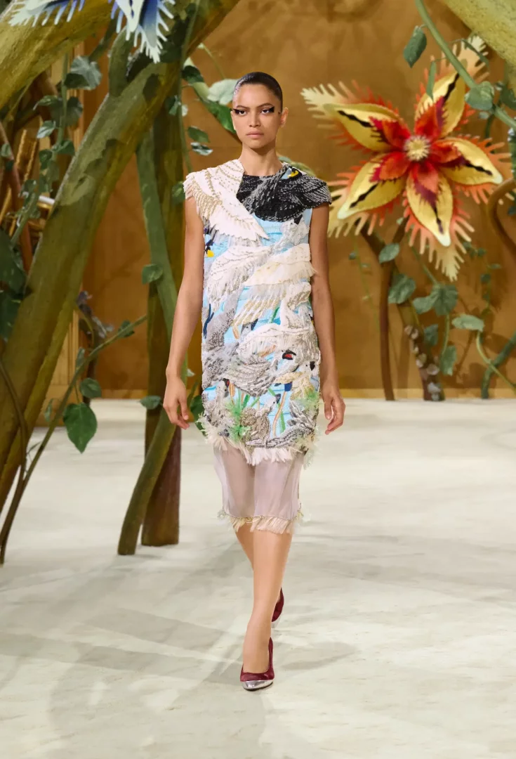 Chanel Sonbahar-Kış 2026 Couture Koleksiyonu'ndan, masallar couture temasını yansıtan, dev çiçekler ve ağaç dallarıyla süslü bir podyumda yürüyen model; kuğu aplikeleri, zengin boncuk ve tüy işlemelerle bezeli, pastel tonlarda kolsuz, katmanlı bir elbise ve bordo topuklu ayakkabılar giyiyor.