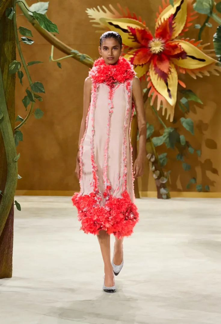 Chanel Sonbahar-Kış 2026 Couture Koleksiyonu defilesinde, masalsı bir podyumda dev çiçeklerin arasında yürüyen model, boynunu ve elbisesinin etek kısmını hacimli mercan rengi tüylerle zenginleştirilmiş, soluk pembe, askılı bir elbiseyle büyüleyici bir şıklık sunuyor.