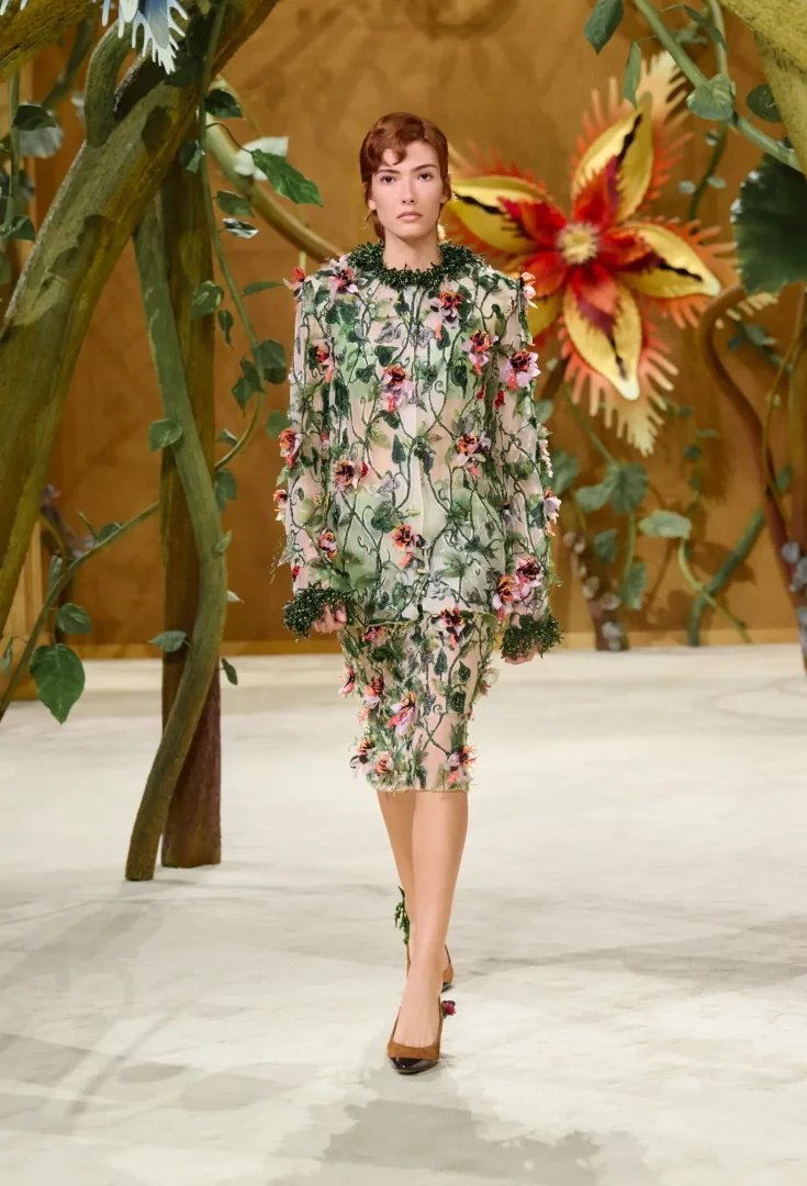Model, Paris Haute Couture Moda Haftası'nda sunulan Chanel Sonbahar-Kış 2026 Couture Koleksiyonu'ndan, yemyeşil dallar ve renkli çiçek aplikeleriyle işlenmiş şeffaf, masalsı bir elbiseyle dev çiçeklerle bezeli bir podyumda yürüyor.