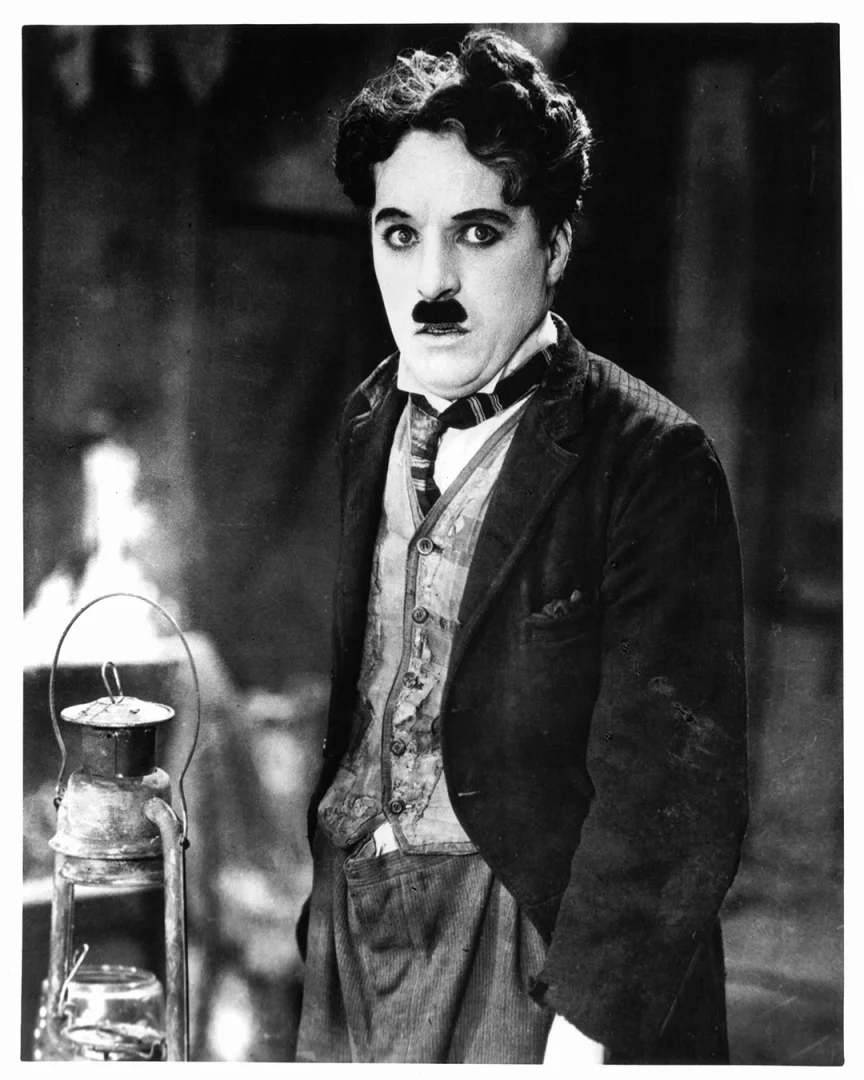Charlie Chaplin'in ikonik Şarlo karakteriyle genişlemiş gözlerle doğrudan izleyiciye baktığı, karakteristik bıyığı ve eski püskü takım elbisesiyle durduğu, sol alt köşede eski bir fenerin görüldüğü bu siyah beyaz fotoğraf, sinema tarihindeki unutulmaz bir anı yakalıyor.