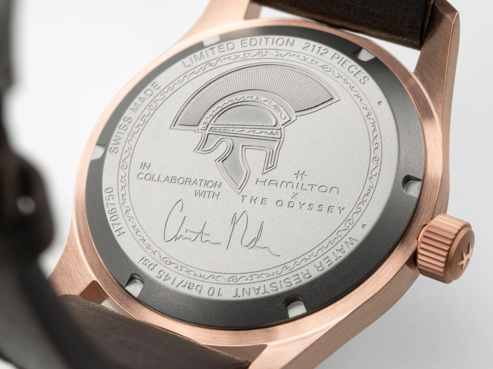 Fırçalanmış rose gold kasalı Hamilton Khaki Field Auto The Odyssey Limited Edition saatinin gümüş renkli arka kapağının yakın çekim görüntüsünde, ortada Spartan miğferi motifinin yanı sıra Christopher Nolan'ın el yazısı imzası, Hamilton ile The Odyssey iş birliğine vurgu yapan yazılar, 2112 adetle sınırlı üretim bilgisi ve 10 bar suya dayanıklılık gibi detaylı işlemeler net bir şekilde yer alıyor.
