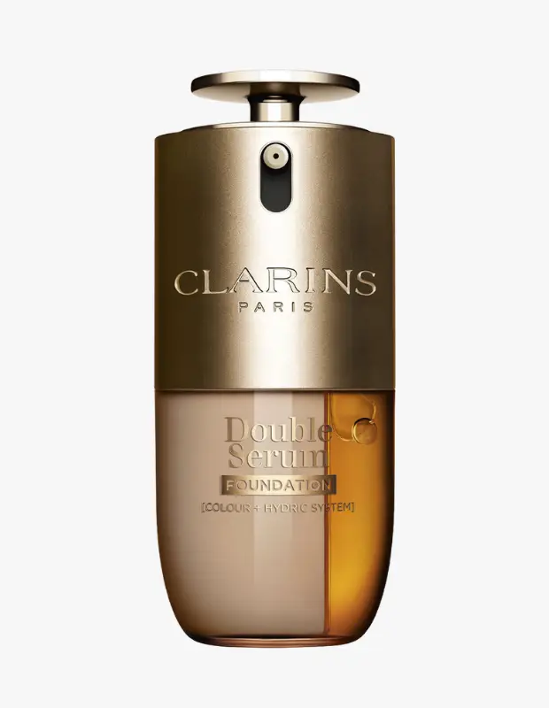 Beyaz bir arka planda, Clarins Double Serum Fondöten şişesi dik duruyor; üst kısmı mat altın rengi pompasıyla birlikte markanın adını taşırken, alt kısmındaki şeffaf bölme ayrık olarak bej fondöten ve parlak kehribar rengi serumu gösteriyor, Colour + Hydric System ibaresi de okunuyor.