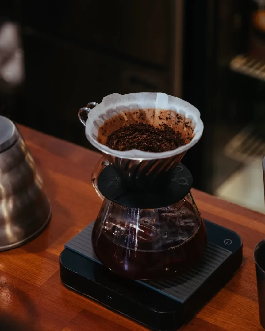 Dijital tartı üzerinde konumlandırılmış cam sürahiye yerleştirilmiş metal bir damlatıcının içindeki beyaz filtre kağıdında, taze demlenmiş kahvenin damla damla toplandığı koyu renkli öğütülmüş kahve taneleri net bir şekilde görülmektedir.