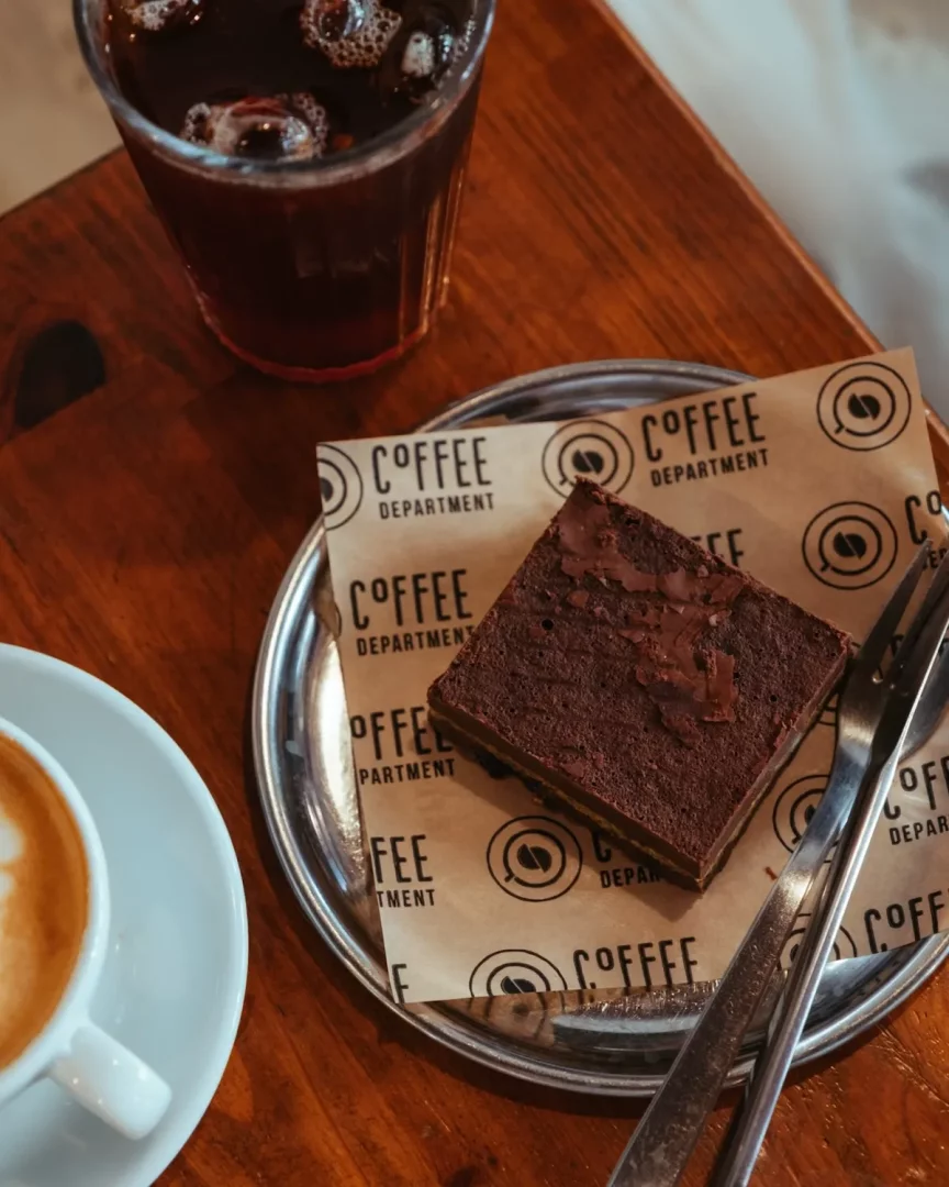 Ahşap bir masada, COFFEE DEPARTMENT markalı kağıt üzerine konmuş parlak metal bir tabakta servis edilen, yanında çatal ve kaşık bulunan bol çikolatalı ıslak brownie, köpüklü sıcak bir latte ve serinletici buzlu kahve, keyifli bir kahve molası için çekici bir sunum oluşturuyor.
