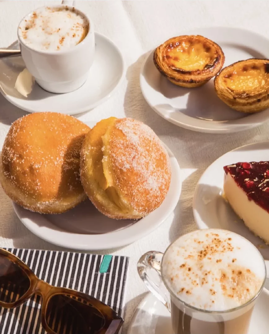 Beyaz bir masada, köpüklü iki kahve, şekerli iki donut, enfes iki Pastel de Nata tart ve kırmızı meyveli bir cheesecake dilimi ile güneş gözlükleri ve çizgili bir defterin eşlik ettiği zengin bir kahvaltı veya tatlı molası görseli.